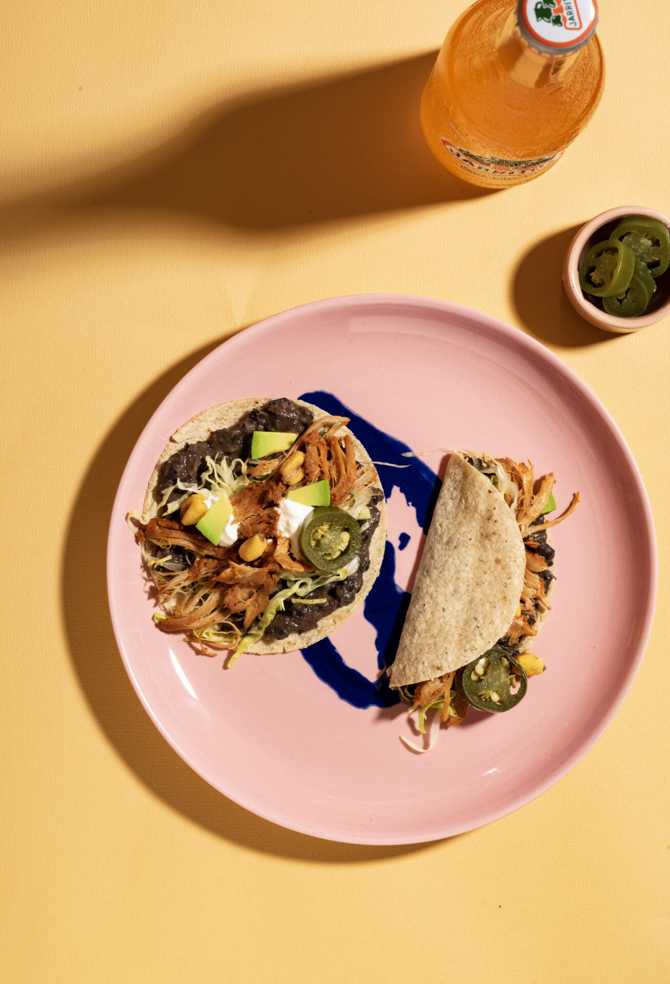 Roze bord met twee mais taco's erop daarnaast staat een flesje Mexicaanse frisdrank